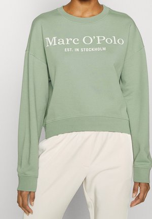 Osoba stojící před neutrálním pozadím má na sobě světle zelenou mikinu s nápisem "Marc O’Polo EST. IN STOCKHOLM" a smetanové kalhoty.