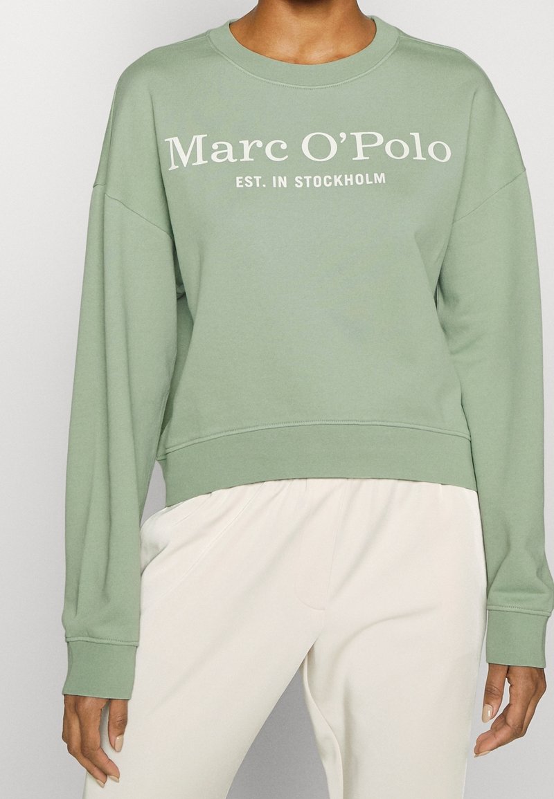 Personne portant un sweat-shirt vert clair avec le texte « Marc O’Polo EST. IN STOCKHOLM » et un pantalon blanc cassé, debout devant un fond neutre.