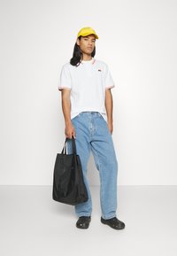 Biała koszulka polo z czerwonymi akcentami, jasnoniebieskie dżinsy, czarna torba, żółta czapka i czarne klapki. Model stoi na jednolitym tle.