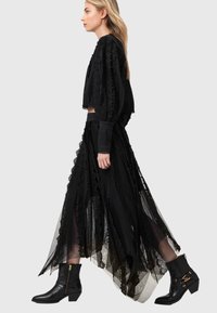 Γυναίκα που περπατάει φορώντας μακριά φούστα από μαύρη δαντέλα και αντίστοιχο μπλουζάκι με λεπτομέρειες, και μαύρα μποτάκια αστραγάλου, πάνω σε απλό φόντο.