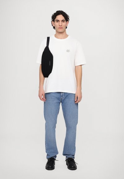 Black crossbody bag worn over a white t-shirt, paired with light blue jeans and black shoes. Model stands against a plain background.
