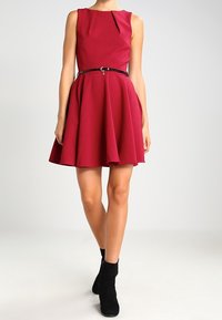 Une robe sans manches, arrivant au genou, de couleur bordeaux, avec un design plissé et une ceinture noire à la taille. Portée avec des bottines noires.