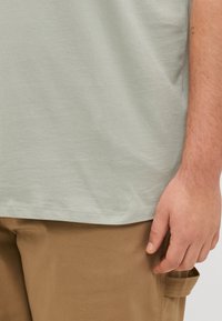 Lys grøn t-skjorte i bomull med rund hals, korte ermer og en avslappet passform, parret med brune bukser med sidelomme.