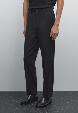 Mann in maßgeschneiderten schwarzen Hosen und schwarzen Lederloafers, der vor einer schlichten grauen Wand und einem dunklen, gesprenkelten Boden steht.