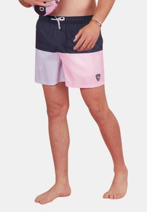 Homme portant un short de bain color-block en rose, marine et lavande, tenant un ballon, debout pieds nus devant un fond uni.