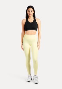 Svart sport-bh och ljusgula leggings, båda åtsittande. Sneakers med vita och grå detaljer. Modellen står upprätt mot en enfärgad bakgrund.