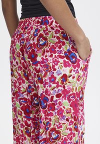 Pantalons à motif floral avec une base rose et des accents rouges, bleus et verts. Taille élastique et poches latérales. Tissu léger avec texture.