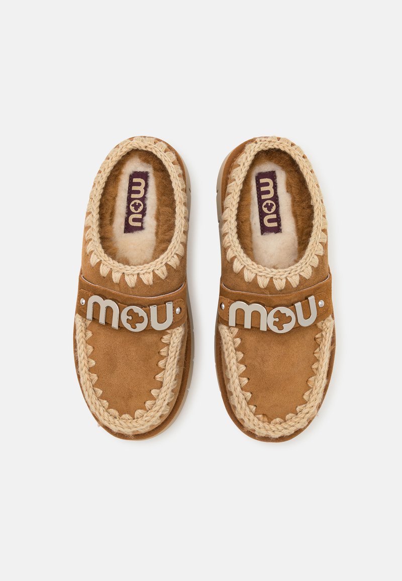 Paire de chaussons mocassins marron avec bordure tricotée crème et doublure intérieure douce, chacun avec une bride portant le logo "mou" sur le dessus.