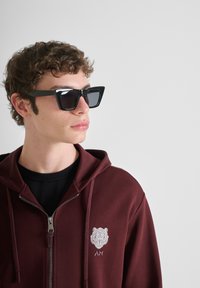 Occhiali da sole neri a forma di occhio di gatto con montatura spessa indossati da una persona in un hoodie bordeaux, con cerniera e logo a forma di leone sul petto.