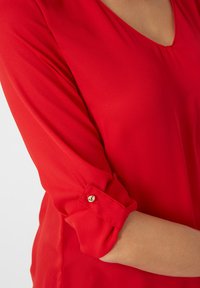 Blusa roja de manga larga con las mangas arremangadas sujetas por un pequeño botón dorado, que muestra parte del brazo y el cuello de quien la lleva.