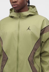 Jeune personne portant une veste à capuche vert clair et marron avec un logo Jumpman noir sur la poitrine et une fermeture éclair à l'avant.