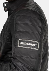 Zwarte leren jas met een doorgestikt ontwerp, voorzien van een grijs geborduurd label "REICHSTADT" op de linkermouw. Zilveren drukknoopsluitingen.