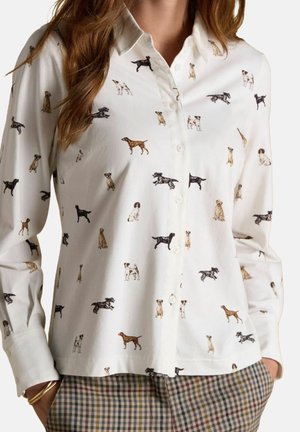 Chemise blanche boutonnée avec un motif de petites illustrations multicolores de chiens, portée avec un pantalon à carreaux, mains dans les poches.