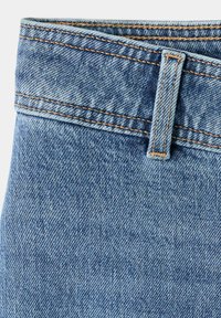 Gros plan de la ceinture d’un jean en denim bleu avec passants de ceinture et surpiqûres orange sur fond clair.