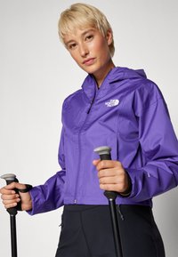 Giacca a vento viola con chiusura a zip, cappuccio e texture liscia. Logo sul petto, mostrata con bastoni da trekking in mano.