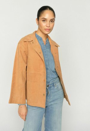 Vrouw draagt een zandkleurige suède jas over een blauw denim overhemd en lichtblauwe spijkerbroek, staand tegen een effen lichtgrijze achtergrond.