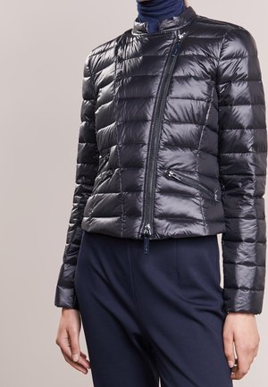 Frau trägt eine glänzende schwarze gesteppte Jacke mit diagonalem Reißverschluss und Reißverschlusstaschen, kombiniert mit dunkler Hose und einem marineblauen Rollkragenpullover.