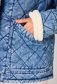 Giacca trapuntata in denim blu sbiadito con un morbido polsino in fleece colore crema. Presenta tasche laterali e dettagli di cucitura blu.