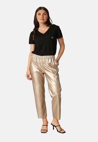 Zwarte V-hals shirt met een klein hartdetail, gecombineerd met glanzende, beige, elastische taillebroek. Model draagt zwarte sleehakken.