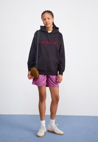 Czarna bluza z kapturem z czerwonym logo, różowe szorty w panterkę, białe skarpetki do kostki oraz kremowe trampki. Brązowa torba na ramię z okrągłym wzorem.