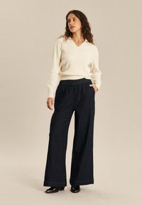 Witte v-hals trui, geribbelde textuur, gecombineerd met losse, donkerblauwe, denim wijde broek en zwarte hoge laarzen, handen in de zakken.