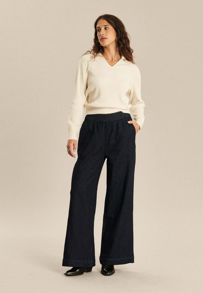 Witte v-hals trui, geribbelde textuur, gecombineerd met losse, donkerblauwe, denim wijde broek en zwarte hoge laarzen, handen in de zakken.