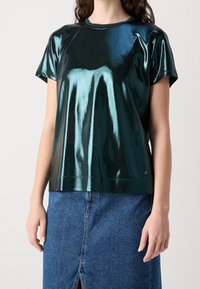 Metallisk grön kortärmad topp med glänsande finish och rundringning, i kombination med en blå denimkjol med en framficka.