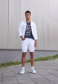 Bijela traper jakna preko tamne majice s grafikom, uparena s bijelim šortsevima i svijetlim tenisicama. Model stoji protiv teksturirane sive pozadine.