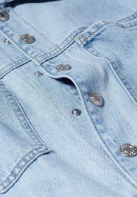 Tissu en denim bleu clair avec une texture douce, présentant des boutons en métal argenté et des détails de couture visibles.