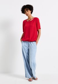 Top rosso a maniche corte con dettagli incrociati sul collo, abbinato a pantaloni in denim a gamba larga di colore blu chiaro. L'outfit presenta una vestibilità comoda e una texture morbida.