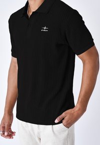 Camisa polo negra con un patrón texturizado de ribetes, mangas cortas, un cuello plano y un logo bordado en blanco en el pecho.