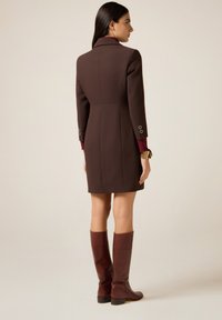 Donna in piedi di profilo che indossa un abito cappotto aderente di colore marrone scuro con maniche lunghe e stivali di pelle marrone fino al ginocchio.