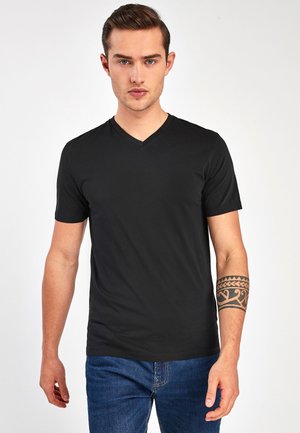T-shirt noir à col en V avec des manches courtes en tissu doux, associé à un jean bleu et arborant un tatouage tribal décoratif sur le bras gauche.