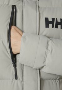 Lichtgrijze pufferjack met een gestructureerde afwerking en zichtbare stiksels. Voorzien van een zwarte ritszak en een opvallend zwart logo op de borst.