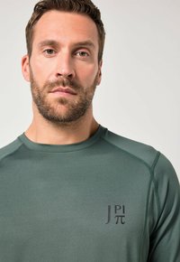 Grøn sportstrøje har en rund halsudskæring, kontrastsyet og et logo trykt på brystet. Stoffet virker glat og fleksibelt.