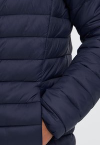 Close-up af en person iført en mørkeblå quiltet jakke med højre hånd i lommen mod en ensfarvet baggrund.
