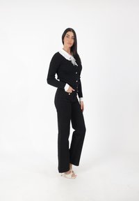 Suéter negro de canalé con un cuello blanco con volantes, combinado con unos vaqueros negros de cintura alta. La modelo lleva unos tacones beige con tiras.