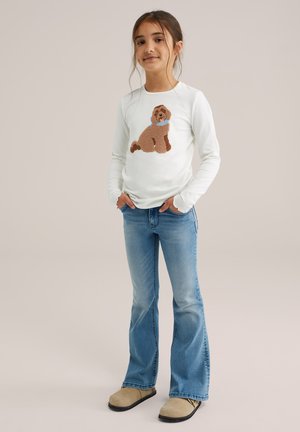 Vit långärmad skjorta med ett strukturerat brunt hundmönster, kombinerad med ljusblå utsvängda jeans och beigefärgade slip-on skor.