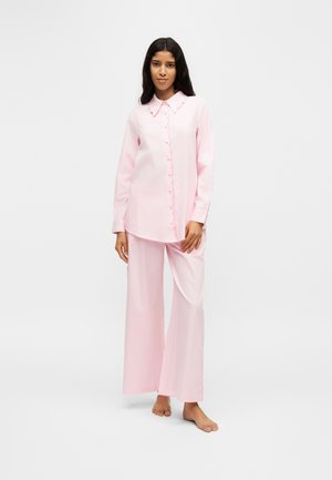 Femme debout pieds nus vêtue d’une chemise rose pâle à manches longues boutonnée et d’un pantalon à jambes larges sur un fond clair uni.