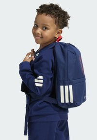 Tmavomodrý batoh s bielymi pruhmi, so zipsom a červeným prvkom. Nosený dieťaťom v zodpovedajúcom tmavomodrom teplákovom odeve s pruhmi.