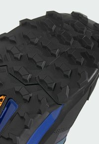 Čierna atletická podrážka s viacerými smerovými gumovými hrotmi, textúrovaným povrchom a oranžovými a modrými akcentmi na boku.