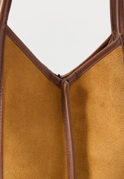 Κοντινό πλάνο γωνίας τσάντας tote από καφέ σουέντ και δέρμα με εμφανείς ραφές και δομημένες ραφές σε απλό φόντο.