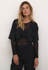 Black textured knit cropped jacket with short sleeves, worn over a sheer black top and paired with black high-waisted trousers.