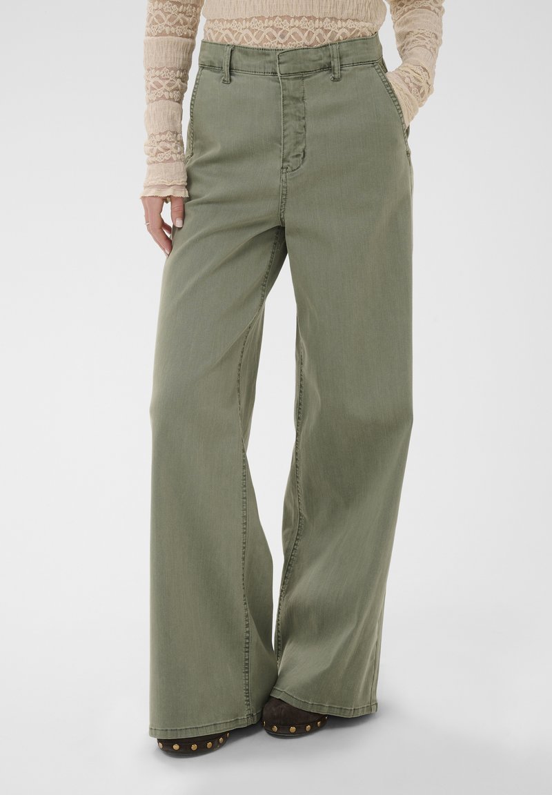 Femme portant un pantalon vert taille haute à jambes larges avec poches avant et un haut beige en dentelle à manches longues, debout avec une main dans la poche.