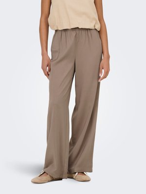 Model iført løse, taupe vide bukser med elastisk talje, kombineret med beige flade sko og en lysebeige ærmeløs top, der er stoppet ind.