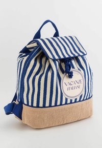 Mochila de lona a rayas en azul marino y crema con un fondo texturizado beige, cierre de solapa y una etiqueta circular que dice "Vacanze Italiane."