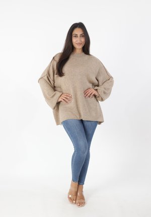Beige oversized gebreid sweater met een hoge halslijn en ballonmouwen, gecombineerd met slim fit blauwe jeans en transparante hakken sandalen.