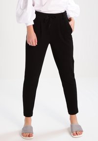 Persoon in zwarte tapse broek met een tailleband met trekkoorden, witte blouse met pofmouwen en grijze open teen slides, staande op een witte vloer.