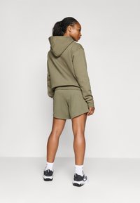 Hoodie court vert olive avec cordon de serrage et shorts assortis, fabriqués en tissu doux ; porté avec des chaussettes blanches et des chaussures de sport noires et blanches.