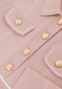 Material tricot roz deschis cu detalii albe cusute. Prezintă două nasturi aurii și buzunare cu clapetă. Textură netedă, elemente de design croite pe măsură.